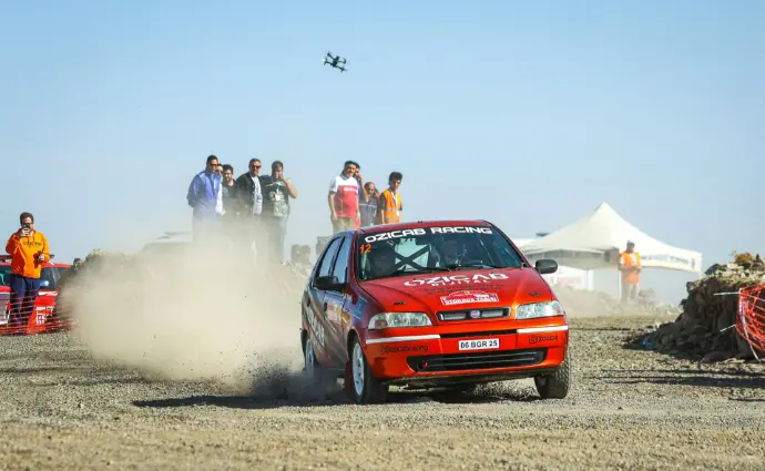 Evento deportivo dron Málaga, grabación aérea rally, fotografía deportiva drones, vídeo carreras Málaga, cobertura eventos dron. Un coche de rally rojo levantando polvo en una pista de tierra durante un evento deportivo, con espectadores observando la acción y un dron volando sobre ellos, capturando la emoción del momento. Esta imagen es perfecta para destacar los servicios de grabación y fotografía aérea con drones para eventos deportivos en Málaga, como rallyes, carreras, maratones o pruebas de ciclismo. Muestra cómo los drones pueden ofrecer perspectivas dinámicas y únicas, mejorando la cobertura y el impacto visual de cualquier acontecimiento deportivo en la provincia.
