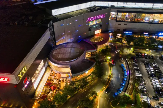 Publicidad dron Málaga, vídeo aéreo centro comercial, marketing digital Málaga, fotografía nocturna dron, promoción negocios Málaga. Impresionante vista aérea nocturna de un gran centro comercial con su estacionamiento lleno de vehículos y las calles circundantes iluminadas, destacando la actividad y el diseño arquitectónico del complejo. Aunque la imagen muestra un "Aeon Mall" que no se encuentra en Málaga, es una excelente representación de los servicios de publicidad y marketing que los drones pueden ofrecer para negocios y centros comerciales en Málaga. Esta fotografía simula cómo se vería un centro comercial en Málaga capital o en la Costa del Sol desde el aire durante la noche, ideal para campañas publicitarias, eventos especiales o la promoción de la vida nocturna y el comercio local.