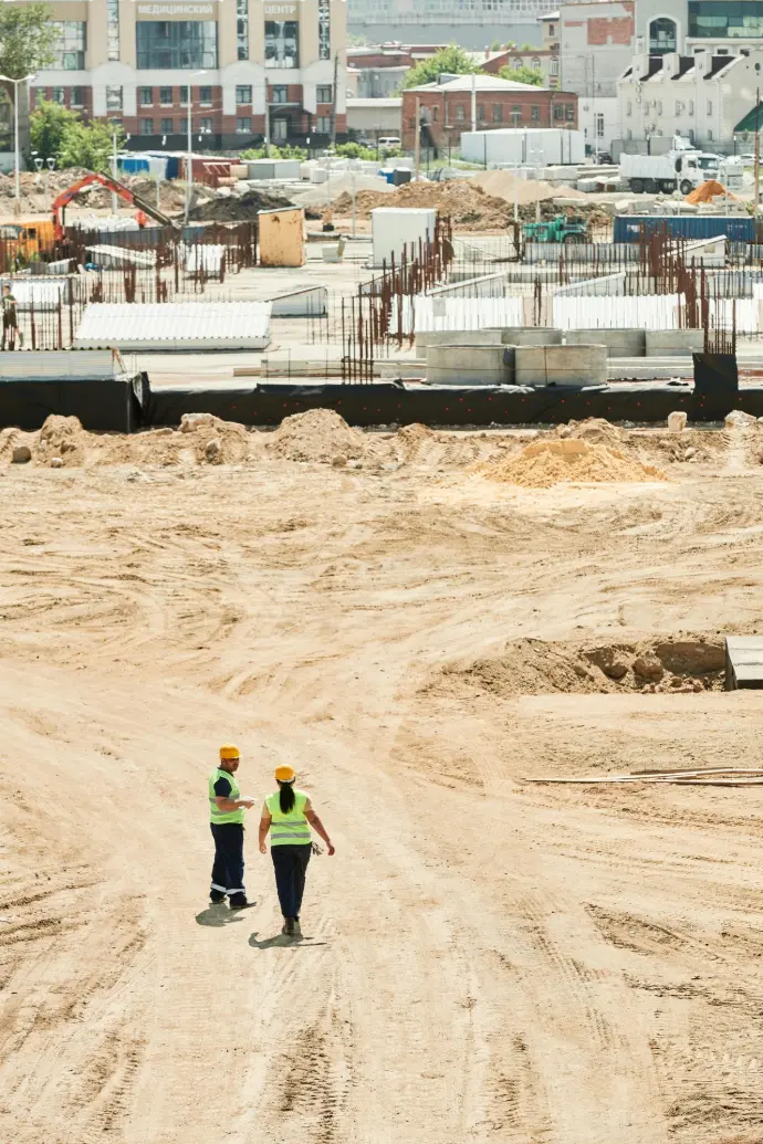 Seguimiento obra Málaga, drones construcción Málaga, inspección aérea obra, avance proyecto dron Málaga, control obras dron. Imagen que muestra a dos trabajadores de la construcción con cascos y chalecos reflectantes inspeccionando una gran obra en Málaga. Se aprecian los cimientos y estructuras en proceso, así como maquinaria pesada al fondo, destacando el progreso del proyecto. Ideal para servicios de seguimiento de obra con drones en Málaga capital y provincia, ofreciendo vistas aéreas precisas para informes de avance.