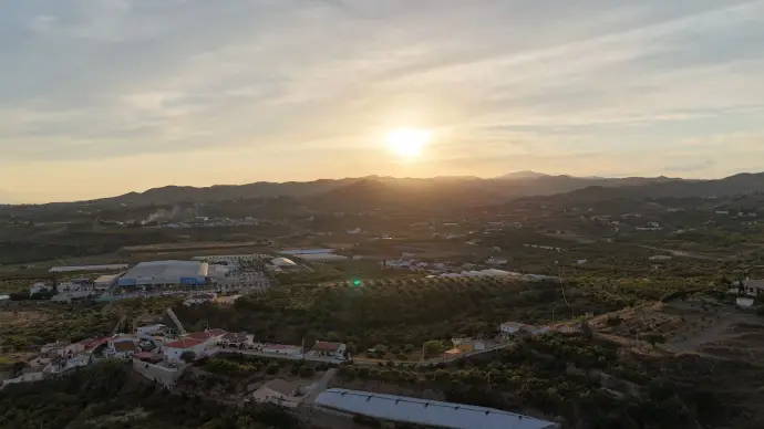 Espectacular vista aérea de un atardecer sobre el paisaje rural de la provincia de Málaga, capturando la luz dorada del sol poniente sobre colinas cubiertas de vegetación, viviendas dispersas y alguna instalación agrícola o industrial. Esta imagen es ideal para mostrar la capacidad de los drones para capturar paisajes impresionantes y la belleza natural de Málaga, ofreciendo servicios de fotografía y vídeo aéreo para promoción turística rural, inmobiliaria en entornos naturales o proyectos audiovisuales en la región.