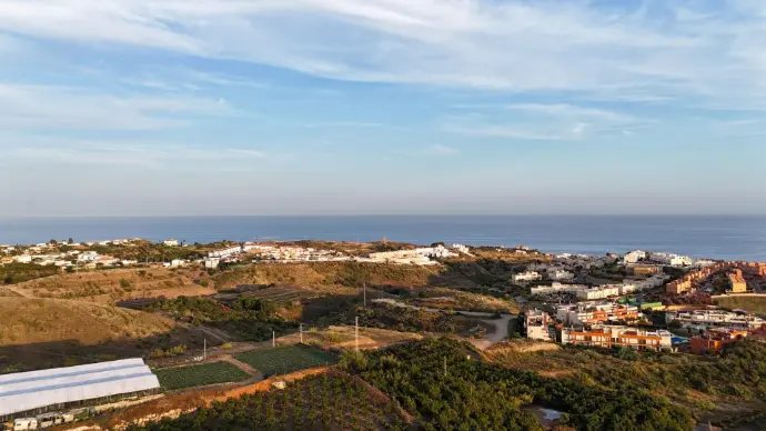 Amplia vista aérea de la costa de Málaga, mostrando la transición de colinas cubiertas de vegetación a zonas residenciales escalonadas que descienden hacia el mar Mediterráneo. Se aprecian viviendas, urbanizaciones y, posiblemente, invernaderos agrícolas en el primer plano, bajo un cielo despejado. Esta imagen es ideal para servicios de fotografía y vídeo aéreo inmobiliario en la Costa del Sol, promoción turística de las playas y pueblos costeros de Málaga, o para proyectos de planificación urbana y seguimiento de desarrollo en la franja litoral malagueña.