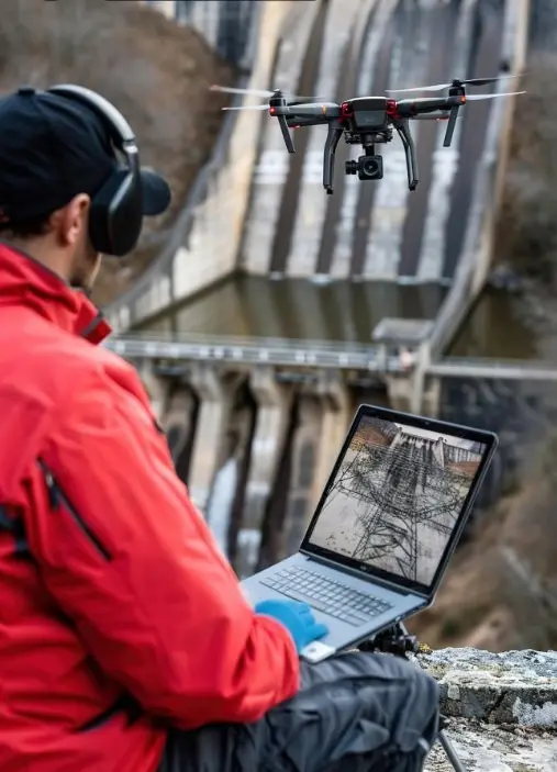 Fotogrametría dron Málaga, inspección embalse, modelado 3D dron, levantamientos topográficos Málaga, dron profesional Málaga. Un técnico profesional opera un dron moderno para realizar trabajos de fotogrametría en un embalse en Málaga, posiblemente para inspección de infraestructura o creación de modelos 3D. Se observa al operador monitoreando la toma en un portátil, con el dron en pleno vuelo y el embalse de fondo. Ideal para empresas y organismos en Málaga que requieran levantamientos topográficos con dron, inspección de presas o modelado 3D de grandes estructuras.