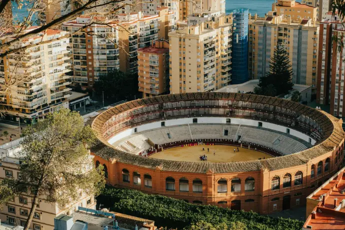 Vista aérea de la Plaza de Toros de La Malagueta en Málaga, mostrando su icónica forma circular y la arena central, rodeada por el denso entramado urbano de edificios y, a lo lejos, el brillo del mar Mediterráneo. Esta imagen resalta la capacidad de los drones para capturar perspectivas únicas de monumentos y áreas urbanas de Málaga, ideal para promoción turística, cobertura de eventos culturales o proyectos inmobiliarios que destaquen la cercanía a puntos de interés en la ciudad.