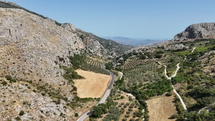 Impresionante vista aérea de un paisaje montañoso en la provincia de Málaga, posiblemente cerca del embalse de La Viñuela, con una carretera serpenteante atravesando el valle, campos de cultivo y olivos, y formaciones rocosas escarpadas. Al fondo, se vislumbran más montañas y una ligera bruma, sugiriendo la proximidad al mar o un vasto horizonte. Esta imagen es perfecta para promocionar servicios de fotografía y vídeo aéreo en entornos naturales de Málaga, ideal para turismo rural, proyectos de ingeniería civil, estudios topográficos o incluso para documentales y producciones audiovisuales que requieran capturar la majestuosidad de la Axarquía y sus paisajes.