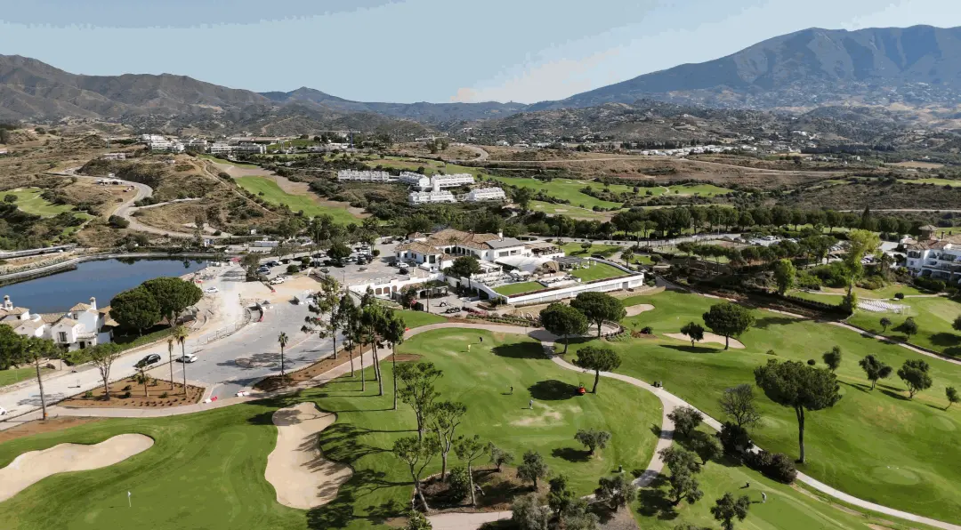 Impresionante vista aérea de un campo de golf de lujo en la Costa del Sol, Málaga, con sus verdes calles, bunkers y un lago artificial, rodeado de complejos residenciales y la majestuosidad de las montañas malagueñas al fondo. Esta imagen es perfecta para destacar los servicios de grabación aérea con drones para promoción inmobiliaria en Málaga, eventos deportivos de golf o incluso inspección de grandes propiedades y complejos turísticos en la región.
