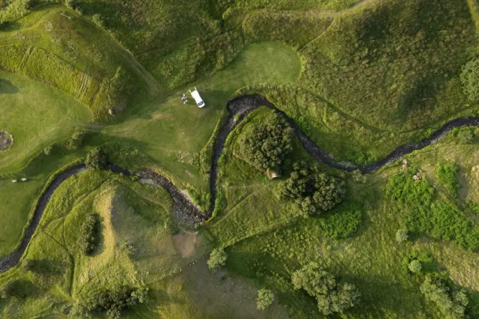 Vista aérea cenital de un hermoso paisaje natural en la provincia de Málaga, mostrando colinas onduladas cubiertas de un verde intenso, un río serpenteante que traza curvas a través del terreno, y un pequeño vehículo tipo autocaravana o furgoneta estacionado en una zona de césped, sugiriendo actividades al aire libre o viajes. La imagen captura la belleza y tranquilidad de los "hermosos paisajes de Málaga" desde una perspectiva única. Es ideal para promover el turismo rural y de aventura en Málaga, servicios de fotografía aérea para campings, fincas rústicas o rutas de senderismo, destacando la riqueza natural de la región.