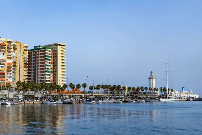 Vista panorámica del puerto de Málaga, mostrando la vibrante combinación de edificios residenciales y hoteleros, palmeras, embarcaciones amarradas en la marina, y el icónico faro de La Farola, con el mar Mediterráneo en primer plano. Esta imagen, aunque nombrada "torremolinos-dron.jpg", representa el puerto de Málaga capital. Es ideal para servicios de fotografía y vídeo aéreo promocional en el ámbito turístico y portuario de Málaga, destacando la belleza de su costa, la infraestructura de su puerto o la vida urbana de la ciudad, perfecta para el sector inmobiliario y de hostelería en la Costa del Sol.
