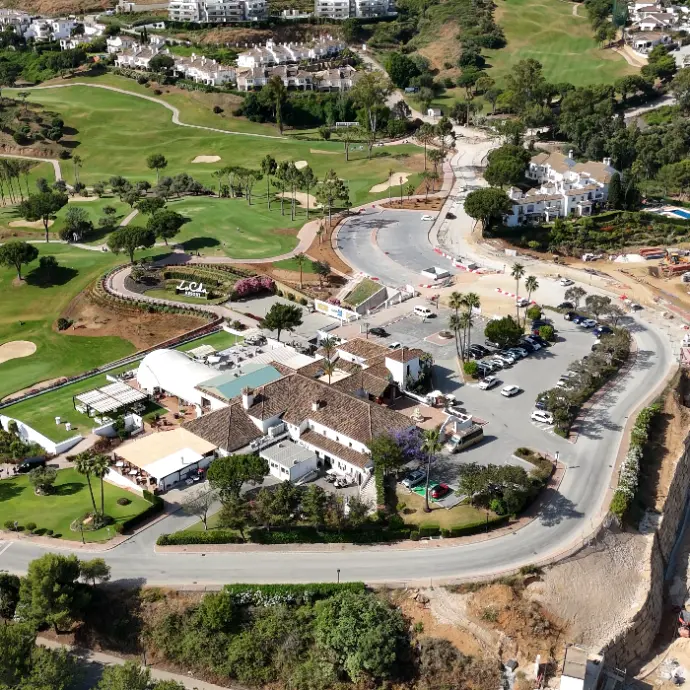 Una vista aérea de un campo de golf y un complejo de edificios, que incluye un club o restaurante, con aparcamientos y carreteras que lo conectan. Se aprecian zonas verdes bien cuidadas y un entorno montañoso con algunas construcciones residenciales al fondo.