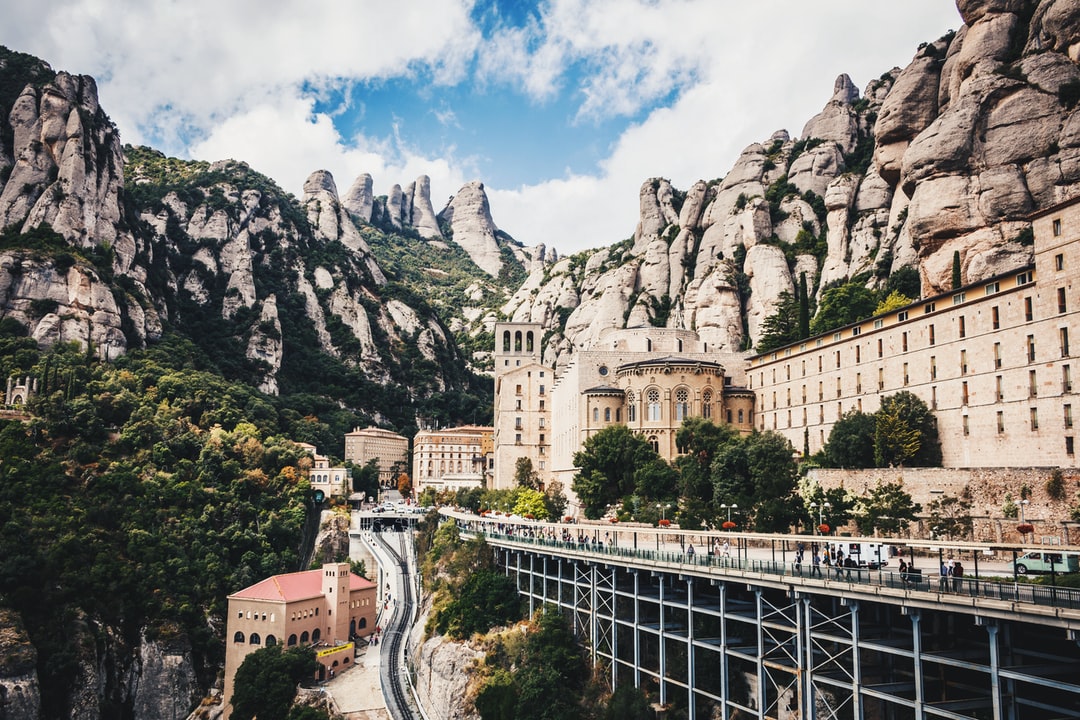Vista panorámica de un impresionante monasterio o complejo de edificios integrado en un paisaje montañoso escarpado, con formaciones rocosas únicas que lo rodean. Una pasarela elevada y una vía de tren serpentean a través del terreno, conectando las diferentes estructuras. Aunque esta imagen no es de Málaga, ilustra perfectamente cómo los drones pueden ofrecer perspectivas espectaculares de lugares de difícil acceso o con gran valor histórico y natural. En el contexto de Málaga, esta técnica sería ideal para capturar la belleza de la Abadía de Santa Ana en Antequera, las formaciones rocosas de El Torcal, o complejos rurales y hoteles boutique enclavados en la Serranía, ofreciendo un servicio único para la promoción turística y cultural de la provincia.